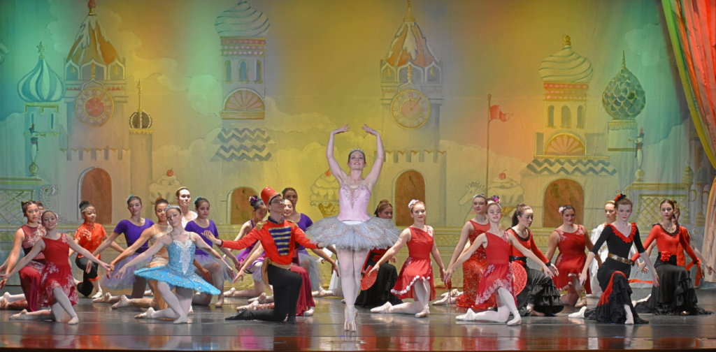 Dancers in various Nutcracker costumes kneel onstage surrounding the Sugar Plum Fairy, who wears a pink tutu.